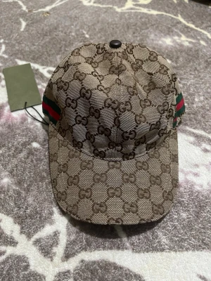 Gucci beige keps med GG-mönster - Snygg beige keps från Gucci med klassiskt GG-monogram över hela kepsen. Kepsen har gröna och röda ränder på sidan och justerbart band bak. Materialet är canvas med insida i vitt tyg. Perfekt för dig som vill ha en exklusiv och trendig accessoar.