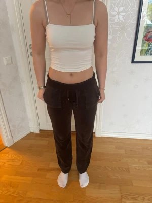 Juicy Couture Bruna mjukisbyxor i velour - Säljer ett par bruna mjukisbyxor i mjuk velour med bred resår i midjan och dragsko. Byxorna har raka ben och stora fickor framtill. Perfekta för en avslappnad och trendig look.