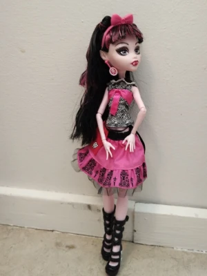 Monster High docka med rosa kläder - Säljer en Monster High docka med långt svart hår och rosa slingor. Dockan har en rosa rosett i håret, svart och vit topp med rosa rosett, samt en rosa kjol med svarta detaljer och spets. Hon har svarta höga skor  Perfekt för samlare eller lek.