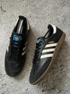 Adidas Samba svarta Og sneaker (Oanvänd) - Klassiska Adidas Samba sneakers i svart läder med vita detaljer och gummisula. Skorna har de ikoniska tre ränderna på sidan, blå logga på plösen och texten 'SAMBA' i guld. Perfekt för dig som gillar retrostil och streetwear. Storlek 44. Snabb Leverans!