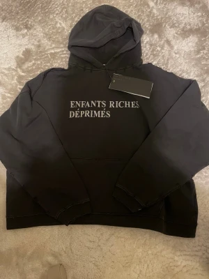 Svart hoodie Enfants Riches Déprimés - Svart hoodie från Enfants Riches Déprimés med vit text på bröstet. Klassisk passform med huva och känguruficka framtill. Tillverkad i mjukt bomullsmaterial och har råa sömmar för en edgy look. Perfekt för dig som gillar streetwear och exklusiva märken.