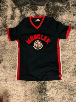 Moncler t-shirt med röd kant - Riktig!!!Snygg mörkblå t-shirt från Moncler med röd kant runt halsen och ärmsluten. T-shirten har vit och röd detalj längs sidorna och stort Moncler-tryck på bröstet. Klassisk v-ringad modell i mjukt material, perfekt för en sportig och trendig look.