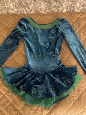 Blå konståkningsklänning  - Unik blå konståkningsklänning med långa ärmar och glittrande blommönster. Klänningen har flera lager med grön tyll under, vilket ger extra volym och en cool kontrast. Figurnära passform. Perfekt för dig som åker konståkning. Köpte för 600 kr.