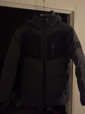 Svart pufferjacka från sail racing - Snygg svart pufferjacka från Tenson med huva och dragkedja framtill. Jackan har vadderad design, två sidofickor och logga på bröstet samt ärmen. Perfekt för kalla vinterdagar och riktigt varm. Materialet är slitstarkt och vattenavvisande.