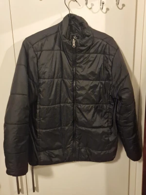 Svart pufferjacka med dragkedja - Snygg svart pufferjacka med hög krage och hel dragkedja framtill. Jackan har ett quiltat mönster och raka ärmar. Materialet ser ut att vara syntetiskt och ger en lätt glansig finish. Perfekt för kalla dagar när du vill hålla värmen och ändå se stilren ut.