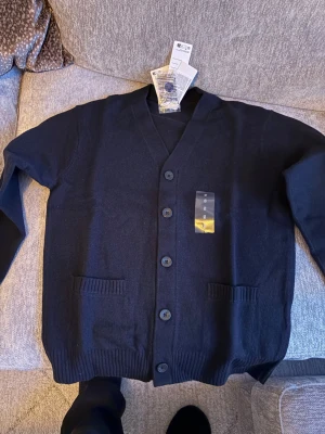 Mörkblå kofta med knappar Uniqlo - Stilren svart kofta från Uniqlo med v-ringning och fem knappar framtill. Två fickor framtill och ribbade muddar vid ärmslut och nederkant. Perfekt att slänga över en t-shirt för en clean look.