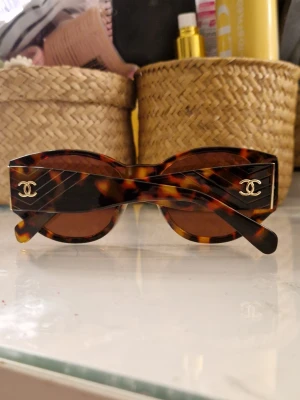Chanel glasögon - Säljer ett par trendiga solglasögon från Chanel med brunmelerad sköldpaddsmönstrad båge i plast. Glasen är bruna och bågarna har den klassiska Chanel-loggan i guld på sidorna. Rektangulär form som ger en cool retrovibe. Perfekt in för sommarn och våren, pris kan diskuteras vid smidig affär