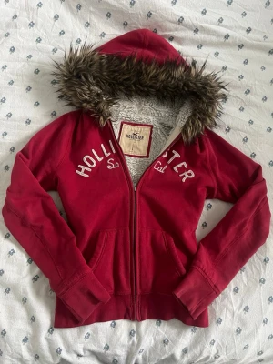 Röd vintage hoodie med fuskpäls från Hollister - Mysig vintage röd hoodie från Hollister med dragkedja, vit brodyrtext på bröstet och stor huva fodrad med fuskpäls. Insidan är klädd i mjuk teddy för extra värme. Perfekt för kalla dagar och chill vibes. Vintage storlek L passar mer som S. måtten finns. 