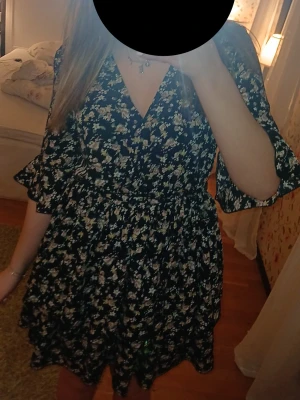 Svart blommig omlottklänning - Kort svart omlottklänning med blommigt mönster i vitt och beige. Klänningen har volangdetaljer på ärmarna, v-ringning och knyt i midjan. Perfekt för sommarens alla tillfällen.