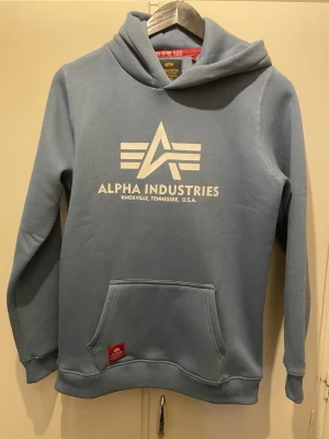 Blå hoodie från Alpha Industries - Snygg ljusblå hoodie från Alpha Industries med stor vit logga och text på bröstet. Klassisk känguruficka framtill och ribbade muddar vid ärmar och nederkant. Tillverkad i mjukt sweatshirtmaterial och har en skön huva. Perfekt för en avslappnad streetwear-look.