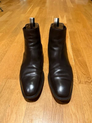 Bruna Boss Chelsea boots i skinn - Stilrena bruna Boss Chelsea boots i slätt skinn med klassisk resår på sidorna och dragflik bak. Snygg, smal siluett och låg klack. Perfekta för en clean och tidlös look.