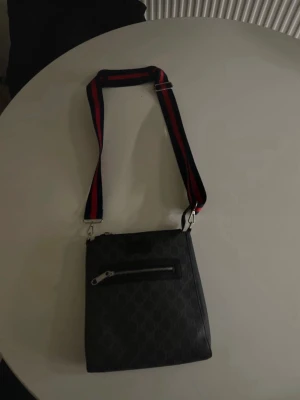 Svart axelväska från Gucci - Snygg svart axelväska från Gucci med diskret GG-mönster och en framficka med dragkedja. Väskan har en bred axelrem i tyg med röda och mörkblå ränder som ger en sportig touch. Perfekt för dig som vill ha både stil och funktion.