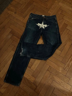 Mörkblå BAPE jeans med vit stjärna - Säljer ett par mörkblå jeans från BAPE med en stor vit blixtformad stjärna tryckt över grenen. Klassisk femficksmodell med raka ben och cool streetwear-vibe. Perfekta för dig som vill sticka ut med en unik detalj på dina jeans. Gissar på ungefär 20 år gamla därför är skicket inte perfekta. Från Japan