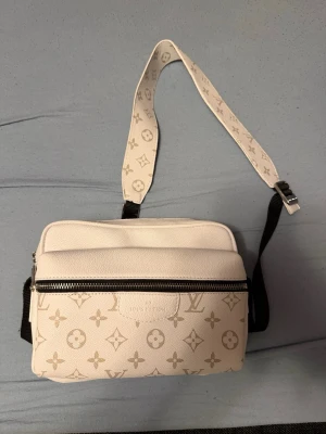  Louis Vuitton väska - Snygg vit axelväska från Louis Vuitton med klassiskt monogrammönster i beige. Väskan har justerbar axelrem med logotyp, dragkedja framtill och är tillverkad i ett tåligt konstlädermaterial. Perfekt för dig som vill ha en trendig och lyxig accessoar.