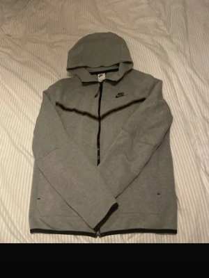 Grå zip hoodie från Nike - Snygg grå hoodie från Nike med dragkedja framtill och svart Nike-logga på bröstet. Hoodien har huva, svarta detaljer längs dragkedjan och nederkanten samt två sidofickor. Perfekt för en avslappnad och sportig stil.