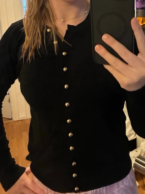 Svart kofta med guldfärgade knappar - Stilren svart kofta med långa ärmar och dekorativa guldfärgade knappar hela vägen framtill. Klassisk rund halsringning och mjukt, tunt material som känns skönt mot huden. Storlek S
