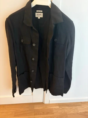 Svart skjorta från East West - Stilren svart skjorta från East West med klassisk krage, knäppning framtill och två bröstfickor. Skjortan har lång ärm och är tillverkad i ett mjukt material som känns bekvämt mot huden. Perfekt för en clean och enkel look.