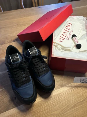Valentino Garavani Rockrunner svarta/blå sneakers - Snygga Rockrunner från Valentino Garavani i svart och mörkblått med coola nitar på sidan. Skorna har en mix av läder och mocka, samt textildetaljer. Klassisk låg modell med svarta skosnören och logga på plösen. Det saknas en nit på insidan av högra skon (se bild) och samma sko har också ett litet märke längst fram vid tårna som inte syns uppifrån. Övrigt i mycket bra skick. Orginalförbackningar, dustbag, nya oanvända skosnören medföljer. Nypris ca 70 00 kr.