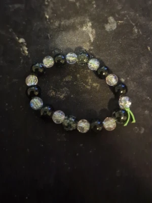 Elastiskt pärlarmband svart och transparent - Snyggt armband med svarta och transparenta pärlor på elastisk tråd. De transparenta pärlorna har en cool, sprucken effekt som ger extra glans. Perfekt accessoar för dig som gillar en unik och stilren look. Tråden är grön vid knuten.