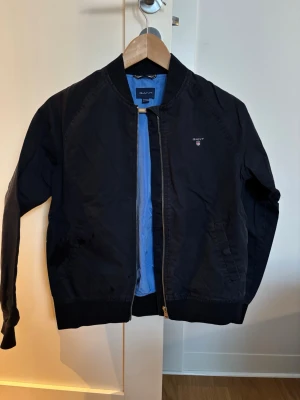 Marinblå bomberjacka från Gant - Snygg marinblå bomberjacka från Gant med klassisk ribbad krage och muddar. Jackan har blått innerfoder, dragkedja framtill och diskret Gant-logga på bröstet. Perfekt för dig som vill ha en stilren och tidlös look.