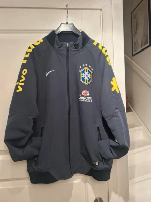 Nike Brasil landslagsträningsoverall - Mörkblå träningsoveralljacka från Nike med Brasilien landslagets emblem på bröstet. Jackan har gula detaljer och tryck på ärmarna, samt sponsorer som Guaraná Antarctica, Vivo och Sadia. Hel dragkedja framtill och två fickor. Perfekt för fotbollsfans.