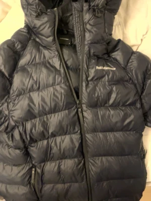 Svart dunjacka från Peak Performance - Snygg svart dunjacka från Peak Performance med huva och dragkedja framtill. Jackan har två sidofickor med dragkedja och diskret logga på bröstet. Perfekt för kalla vinterdagar tack vare det tjocka dunfodret och den höga kragen.