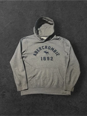 Grå hoodie från Abercrombie & Fitch - Snygg grå hoodie från Abercrombie & Fitch med mörkblå text och logga framtill. Klassisk känguruficka och huva. Tillverkad i mjukt bomullsmaterial som känns skönt mot huden. Perfekt för en avslappnad och trendig look.