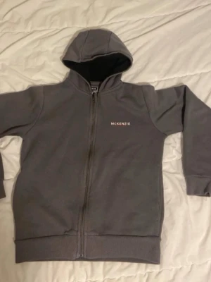 Grå zip hoodie från McKenzie - Säljer en stilren grå hoodie från McKenzie med dragkedja framtill och huva. Hoodien har diskret logga på bröstet och ribbade muddar vid ärmar och nederkant. Perfekt för chill dagar eller när du vill ha en enkel look.