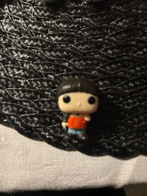 Will Stranger things kinder joy - En liten samlarfigur i plast med svart hår, stora ögon och röd tröja. Figuren har blå byxor och svarta skor, och är designad i en söt, tecknad stil. Perfekt som dekoration eller till samlingen.