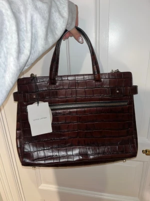  Little Liffner modell Funky b-l-chocola väska - Snygg mörkbrun handväska från Little Liffner i croco-präglat läder. modell: Funky b-l-chocola  Väskan har både handtag och avtagbar axelrem i silverfärgade detaljer samt en stor dragkedjeficka framtill. Prislappen kvar den är oanvänd. Nypris är 5400. Skriv om du har frågor eller vill ha fler bilder💕