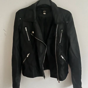 Svart skinnjacka från ONLY - Snygg svart skinnjacka från ONLY med klassisk bikerstil, sneddragna silverfärgade dragkedjor och flera fickor. Jackan har bred krage och detaljerade sömmar på axlarna. Perfekt för en cool och trendig look.
