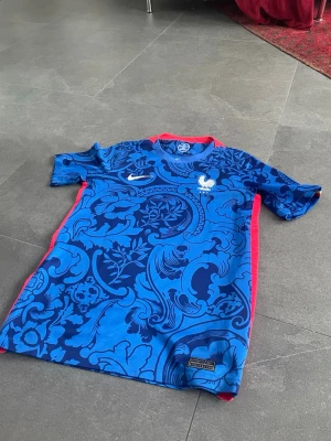 Blå Frankrike matchtröja Nike - Säljer en blå matchtröja från Nike för Frankrikes landslag. Tröjan har ett unikt blommigt mönster i olika blå nyanser, röda detaljer längs sidorna och runt kragen samt Frankrikes tupp-logga och Nike swoosh på bröstet. Perfekt för en clean look passar skit bra till ett par jeans!! Priset är inte hugget i sten!!