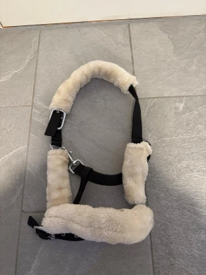 Svart grimma med beige fuskpäls - Säljer en svart grimma med mjuk beige fuskpäls på insidan. Grimman har justerbara spännen i metall och passar perfekt till häst eller ponny. Materialet är slitstarkt nylon med fluffig fuskpäls för extra komfort.