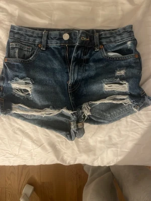 Blå slitna jeansshorts med hög midja - Snygga blå jeansshorts med hög midja och slitna detaljer framtill och baktill. Klassisk femficksmodell med knapp och dragkedja. Perfekta för en avslappnad stil under varma dagar.