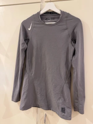 Nike PRO thermal dri-fit tröja  - Snygg grå långärmad träningströja från Nike Pro med diskret struktur i tyget och vit logga på axeln. Tröjan har rund halsringning och är tillverkad i ett stretchigt funktionsmaterial som andas, perfekt för träning och sport.