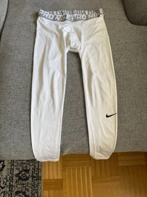 Vita Nike Pro träningstights - Säljer ett par vita Nike Pro träningstights med bred resår i midjan där det står 'NIKE PRO' i grått. Tightsen har en tight passform och är gjorda i ett stretchigt funktionsmaterial. Svart Nike-logga på vänster ben.