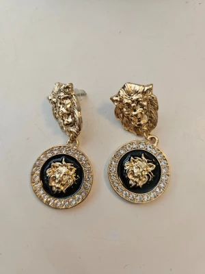 Guldiga örhängen med lejonmotiv - Snygga örhängen i guldfärgad metall med lejonhuvud som detalj. Ena hänget har svart bakgrund och gnistrande stenar runt kanten, vilket ger en lyxig känsla. Perfekt accessoar för att sticka ut och visa attityd.