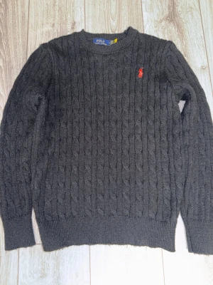 Mörkgrå kabelstickad tröja Polo - Kabelstickad mörkgrå tröja från Polo Ralph Lauren med rund halsringning och röd broderad logga på bröstet. Tröjan har ribbade muddar vid ärmslut och nederkant, vilket ger en klassisk och stilren look. Perfekt för dig som gillar tidlös design.