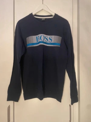Mörkblå sweatshirt från BOSS - Mörkblå sweatshirt från BOSS med stort logotryck i blått och vitt framtill. Klassisk rund halsringning och långa ärmar. Perfekt för en avslappnad och stilren look.