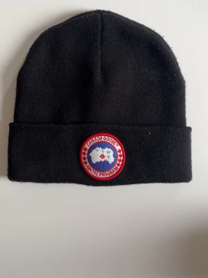 Svart mössa från Canada Goose - Snygg svart mössa från Canada Goose med klassisk uppvikt kant och det ikoniska runda röda 