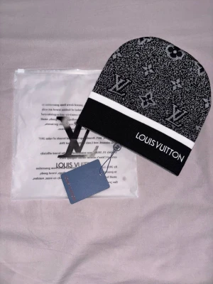 Louis Vuitton svartvit mössa - Snygg svartvit mössa från Louis Vuitton med klassiskt LV-monogram och logotyp framtill. Mössan har en vit rand och är tillverkad i mjukt stickat material. Perfekt accessoar för dig som vill ha en exklusiv och trendig look.