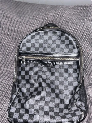 Svart och grå rutmönstrad ryggsäck - Snygg ryggsäck med svart och grått rutmönster, dragkedja framtill och justerbara axelband. Väskan har en klassisk form och är tillverkad i ett tåligt material med detaljer i konstläder och mesh på sidorna. Perfekt för dig som gillar stilrena accessoarer.