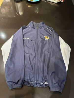 Blå vindjacka från CCM TG - Snygg blå vindjacka från CCM med TG-logga och vita detaljer på ärmarna. Jackan har dragkedja framtill, två sidofickor och resår i midja och ärmslut. Perfekt för dig som gillar sportig stil och vill ha en lätt jacka till vardagen.