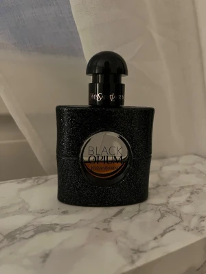 Yves Saint Laurent Black Opium parfym - Black Opium från Yves Saint Laurent är en ikonisk parfym i en svart, glittrig flaska med rundad form och transparent cirkel i mitten. Flaskan har en elegant svart kork och tydlig logga framtill.
