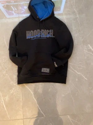 Svart hoodie från Hoodrich med blå huva - Svart hoodie från Hoodrich med stor logga i vitt och blått på bröstet, blå insida i huvan och dragsko. Magficka framtill och patch med text på fickan. Ärmen har en vit broderad detalj. Perfekt för dig som gillar streetwear och snygga detaljer.