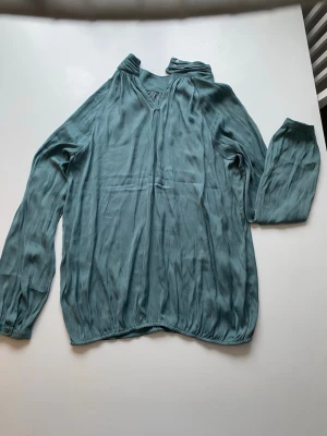 Blågrön blus med v-ringning - Snygg blågrön blus i ett tunt, mjukt tyg med v-ringning och lätt rynkad krage. Blusen har långa ärmar med knapp vid ärmslut och avslappnad passform. Perfekt att styla med jeans eller kjol för en trendig look.