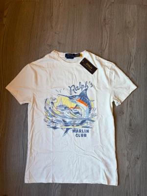 INTRESSEKOLL  - En sällsynt vit t-shirt från Polo Ralph Lauren med tryck av texten 'Ralph's Marlin Club' på framsidan. Klassisk rund hals och kort ärm. Classic fit-modell, perfekt för en avslappnad stil. Nypris: 1200, skriv om ni har prisförslag! Självklart äkta✅