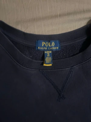 Mörkblå sweatshirt från Polo Ralph Lauren - Klassisk mörkblå sweatshirt från Polo Ralph Lauren med rund halsringning och röd broderad logga på bröstet. Tröjan har långa ärmar och ribbade muddar vid ärmslut och nederkant. Perfekt för en avslappnad och stilren look.
