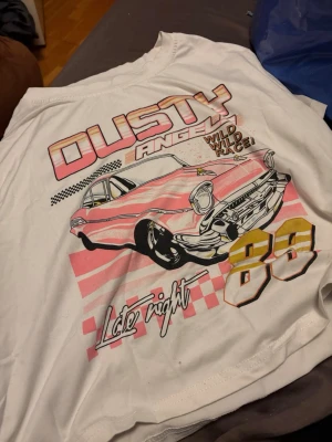 Vit t-shirt med biltryck och text - Snygg vit t-shirt med stort retroinspirerat tryck av en rosa bil, texten 'DUSTY ANGEL', 'Wild Race' och siffran 88 i gult. T-shirten har korta ärmar och cool streetstyle-vibe, perfekt för dig som gillar unika prints.