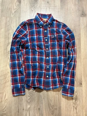 Hollister - Riktigt snygg rutig skjorta från Hollister i blått, rött och vitt. Skjortan har klassisk krage, knappar framtill och lång ärm. Perfekt för dig som gillar en avslappnad men ändå stilren look.